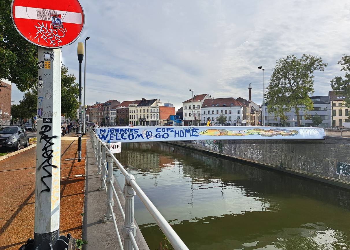 13 september 2022: een nieuw opschrijft op de passerelle over het kanaal ter hoogte van het MIMA:  "Migrants welcom(e), Cops go home"