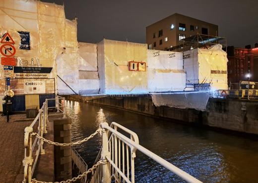 De voor renovatie met krimpfolie ingepakte historische Godshuizenbrug over het kanaal kreeg van een kunstliefhebber een paneel dat de voorbijganger vraagt even stil te staan bij de brug en het werk van het kunstenaarskoppel Christo Vladimiroff Javacheff (1935-2020) en Jeanne-Claude Denat de Guillebon (1935-2009). Dit duo verwierf wereldwijd bekendheid met het inpakken van hitorische gebouwen zoals de Reichstag in Berlijn in 1995 en de Pont Neuf in Parijs in 1985.